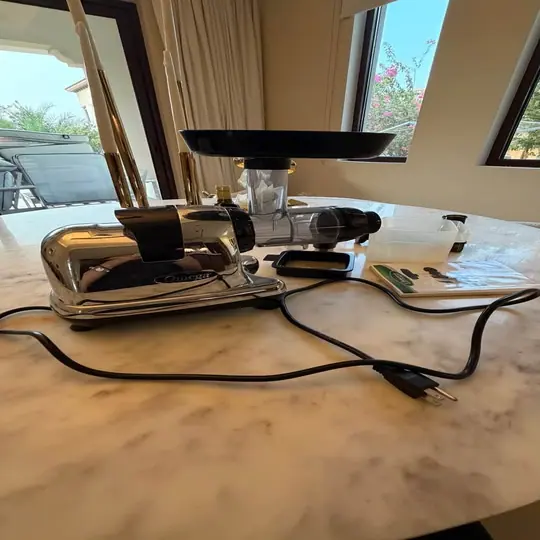 Chrome masticating juicer with its accessories laid out on a marble table
